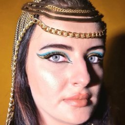 Renata Czapska - Portret kobiety z makijażem inspirowanym stylem egipskim: turkusowe kreski, złote akcenty i ozdobny, złoty łańcuch na głowie na tle żółtej ściany.