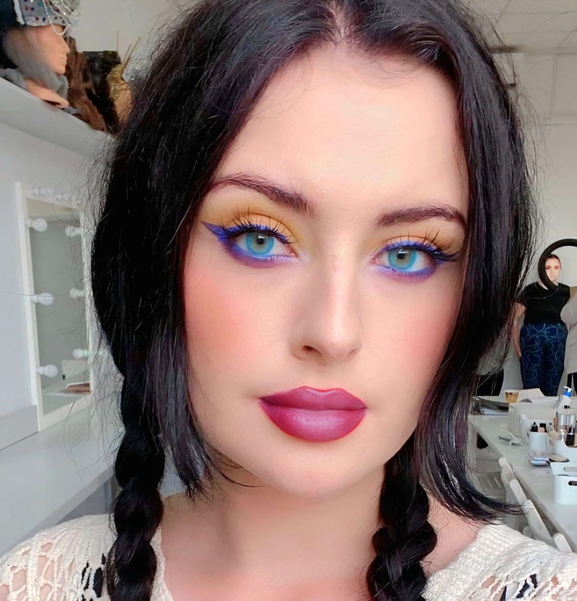 Portret kobiety z wyrazistym, artystycznym wizażem: niebieskie oczy, żółto-złote cienie i ciemnoróżowe usta. Włosy zaplecione w dwa warkocze, bluzka z koronki.