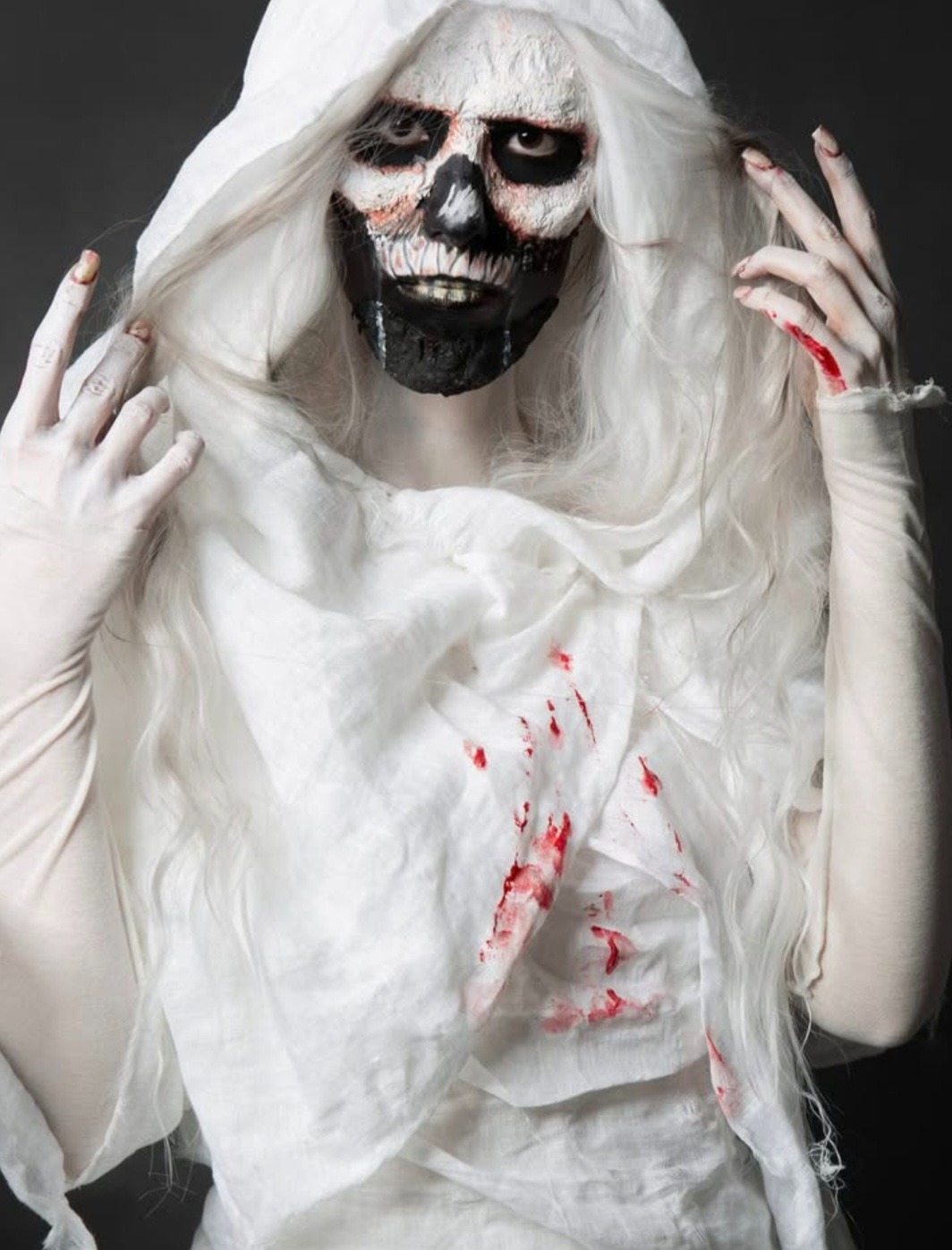 Osoba w kostiumie ducha z makabrycznym efektem czaszki i plamami imitującymi krew na białym przebraniu, z wyciągniętymi dłońmi na ciemnym tle.