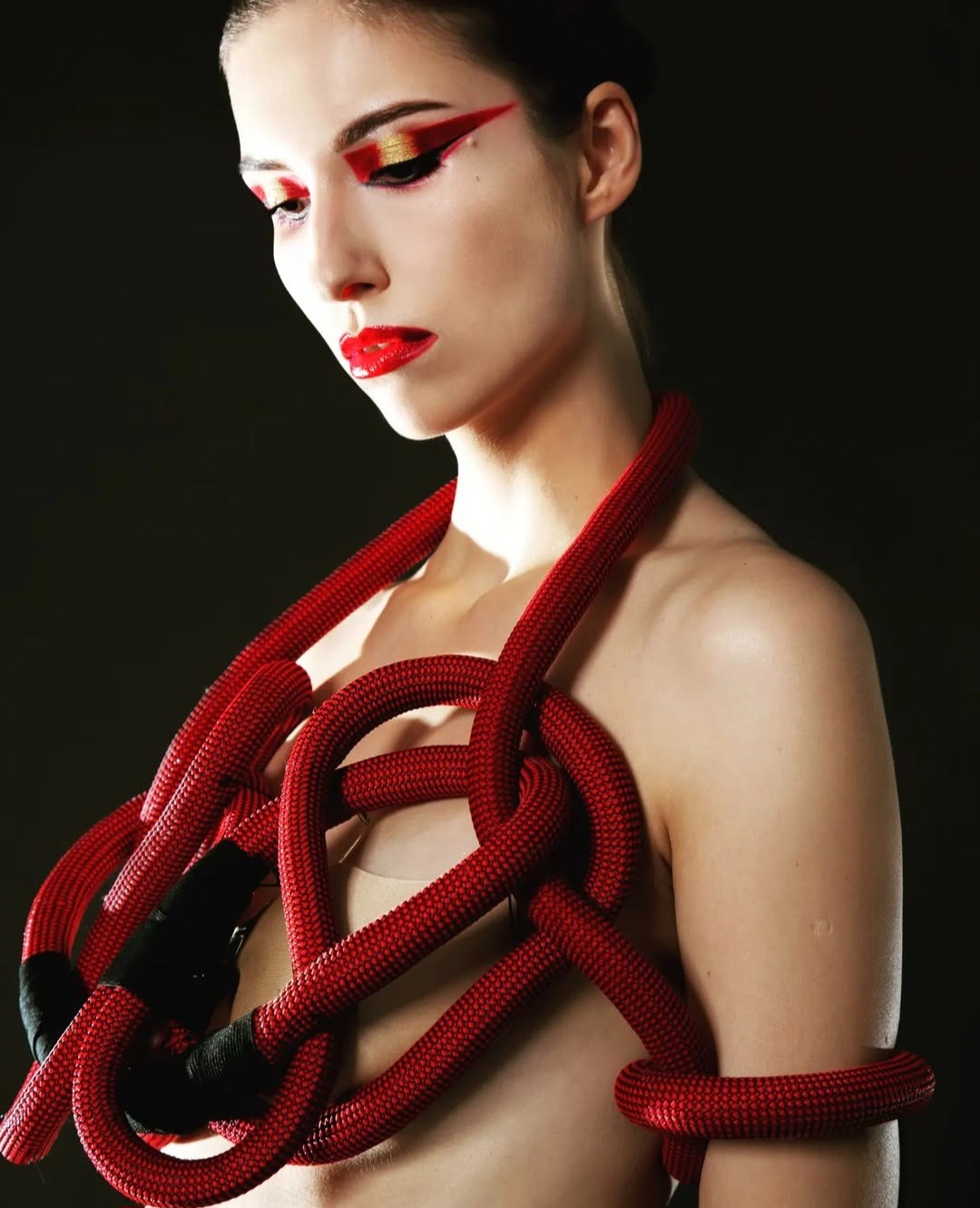Portret kobiety z awangardowym wizażem: czerwone usta i graficzny makijaż oczu w kolorach czerwieni i złota. Uwagę przykuwa nietypowa biżuteria z czerwonej liny.