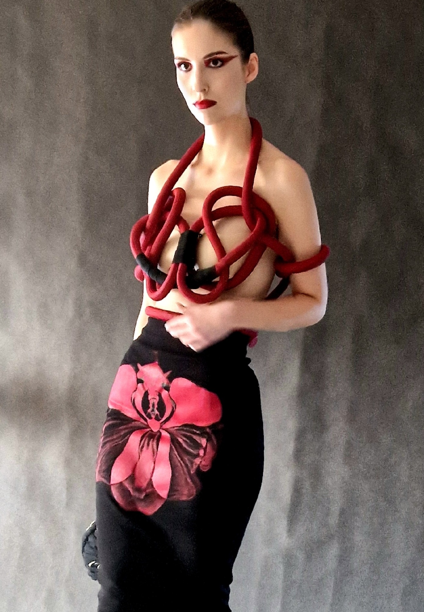 Modelka z intensywnym makijażem oczu i ust w stylizacji artystycznej z czerwonymi elementami, na szarym tle. Wyrazisty make-up i unikalny strój.