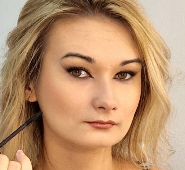 Portret kobiety z blond włosami i wyraźnym makijażem oczu typu kocie oko, podkreślone usta i delikatny róż na policzkach. W kadrze dłoń z pędzlem.