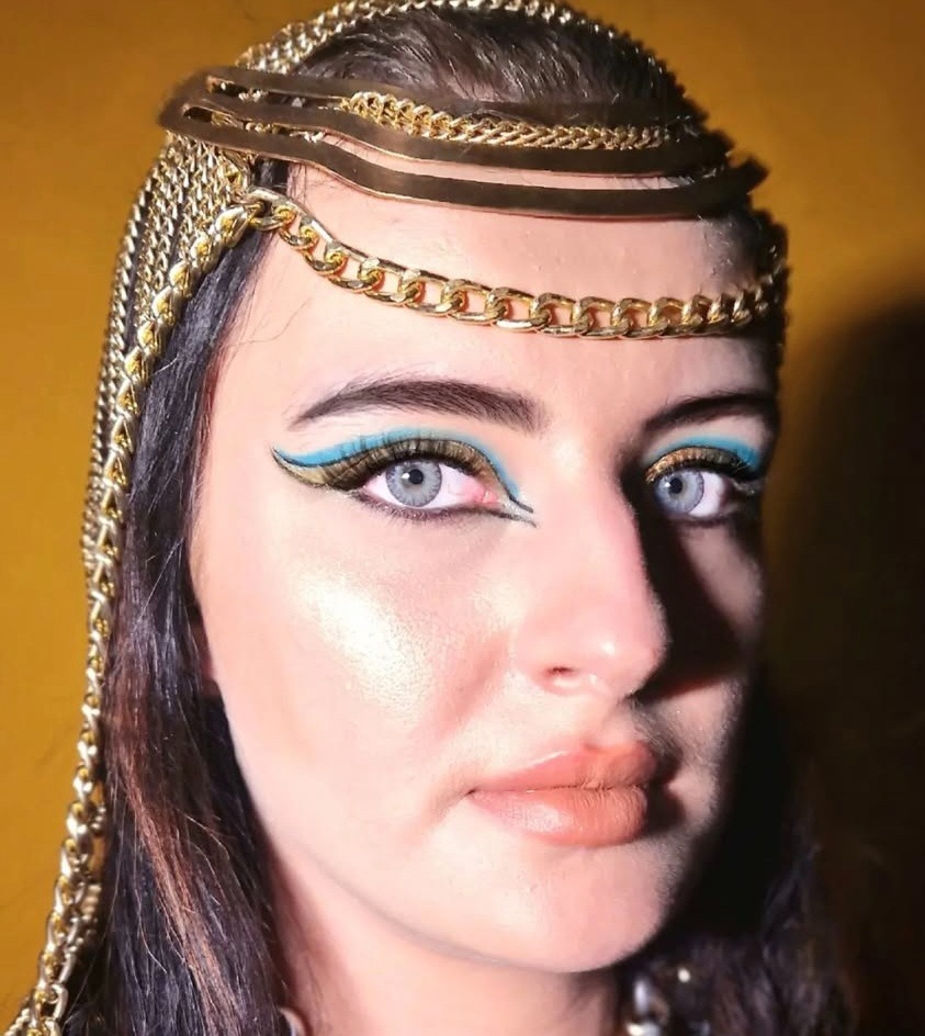 Portret kobiety z makijażem inspirowanym stylem egipskim: turkusowe kreski, złote akcenty i ozdobny, złoty łańcuch na głowie na tle żółtej ściany.