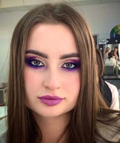 Portret kobiety z profesjonalnym makijażem: fioletowe cienie i matowa pomadka. W tle widoczne stanowisko kosmetyczne i inne osoby.