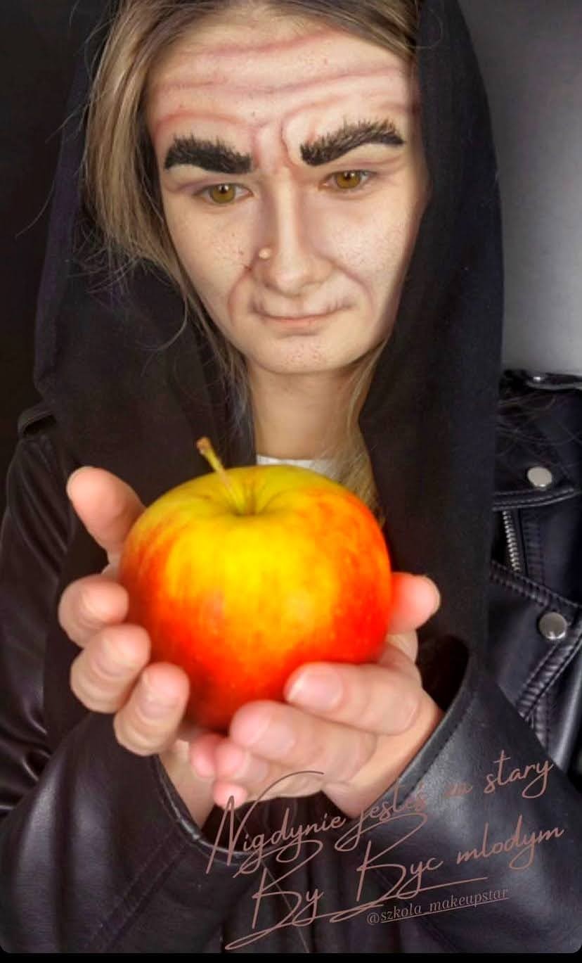 Osoba z charakteryzacją na starą wiedźmę, trzymająca jabłko w dłoniach. Widoczne zmarszczki i brwi podkreślone charakteryzacją. Ubrana w czarną bluzę i skórzaną kurtkę.