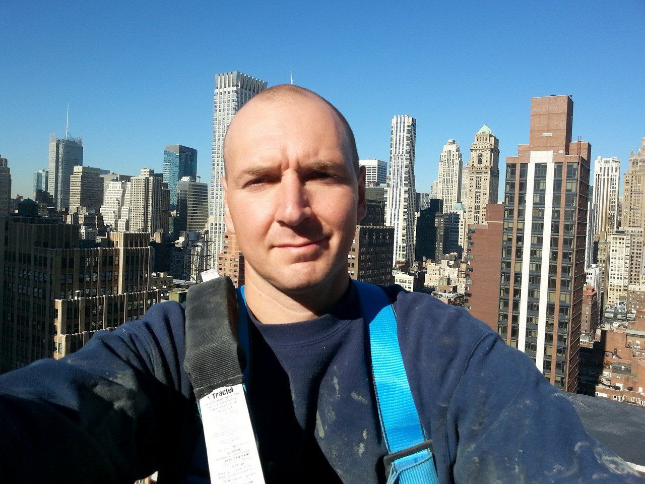 Mężczyzna w uprzęży na dachu wieżowca, panorama Manhattanu w tle. Ujęcie z dołu, niebieskie niebo, widoczne budynki w różnych stylach architektonicznych.