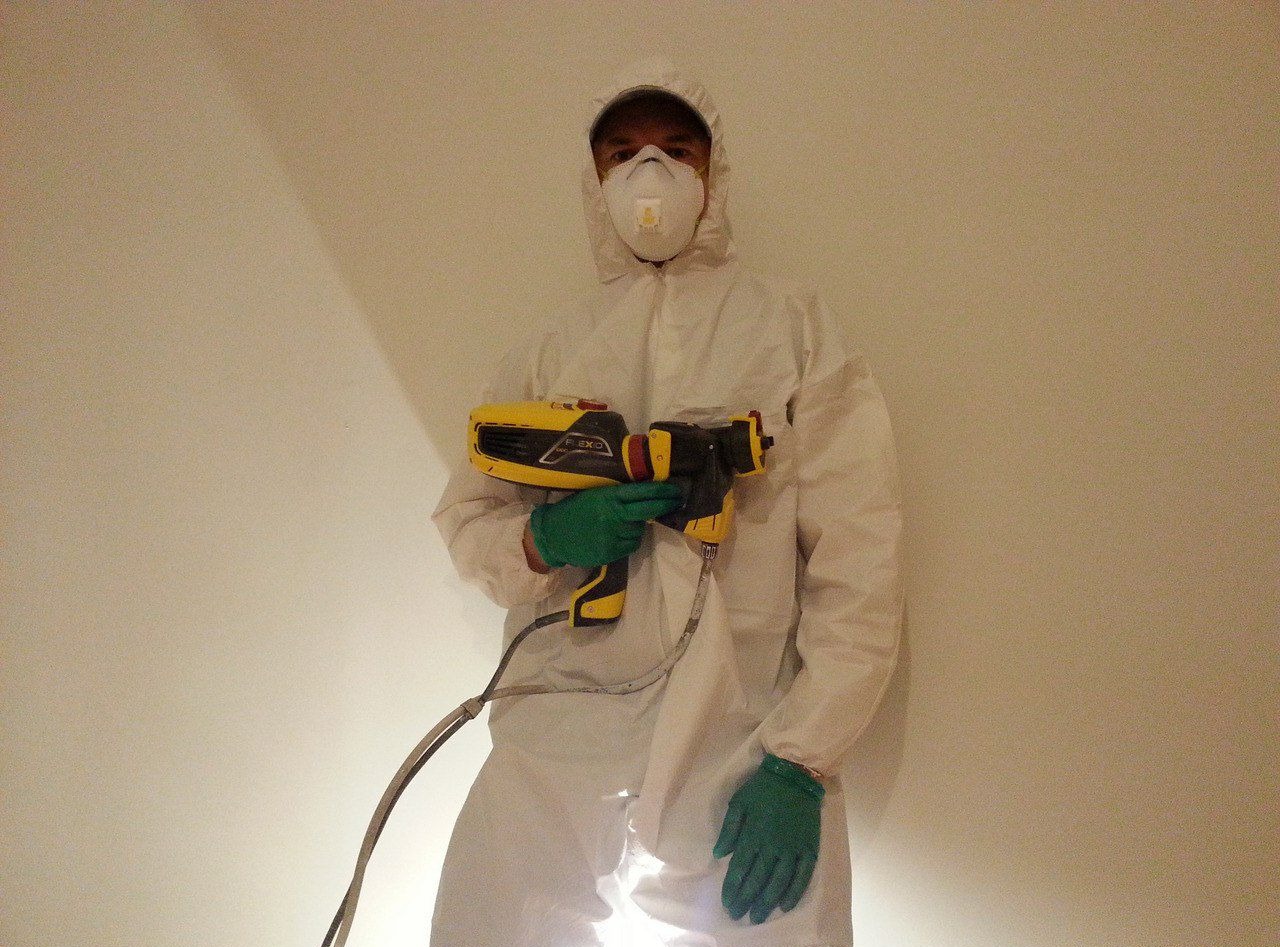 Pracownik w kombinezonie ochronnym i masce trzyma pistolet natryskowy Flexio na tle jasnej ściany. Widoczne zielone rękawice i przewód urządzenia.