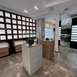 Wnętrze salonu optycznego: ekspozycja okularów w podświetlanych gablotach na ścianie i w szklanych witrynach na postumentach. Nowoczesny design, jasne oświetlenie.