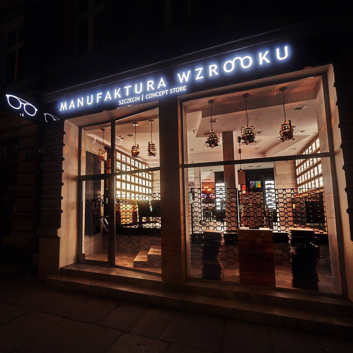 Nocna fasada salonu optycznego 'Manufaktura Wzroku' w Szczecinie. Podświetlony szyld, wystawa okularów i dekoracyjne lampy wiszące.
