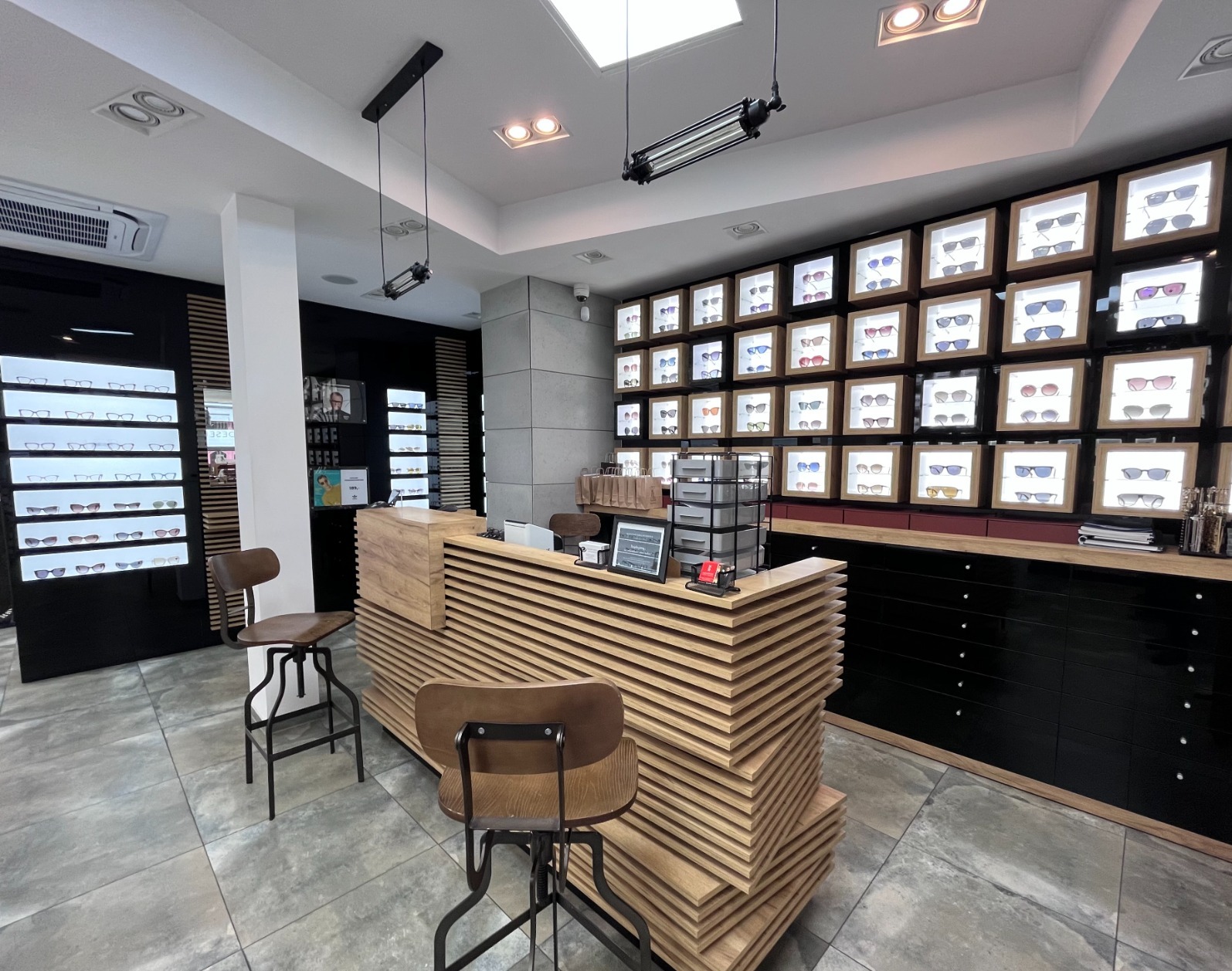 Nowoczesny salon optyczny: drewniana lada recepcyjna z ekspozycją okularów w podświetlanych gablotach. Minimalistyczny design, ciepłe oświetlenie punktowe.