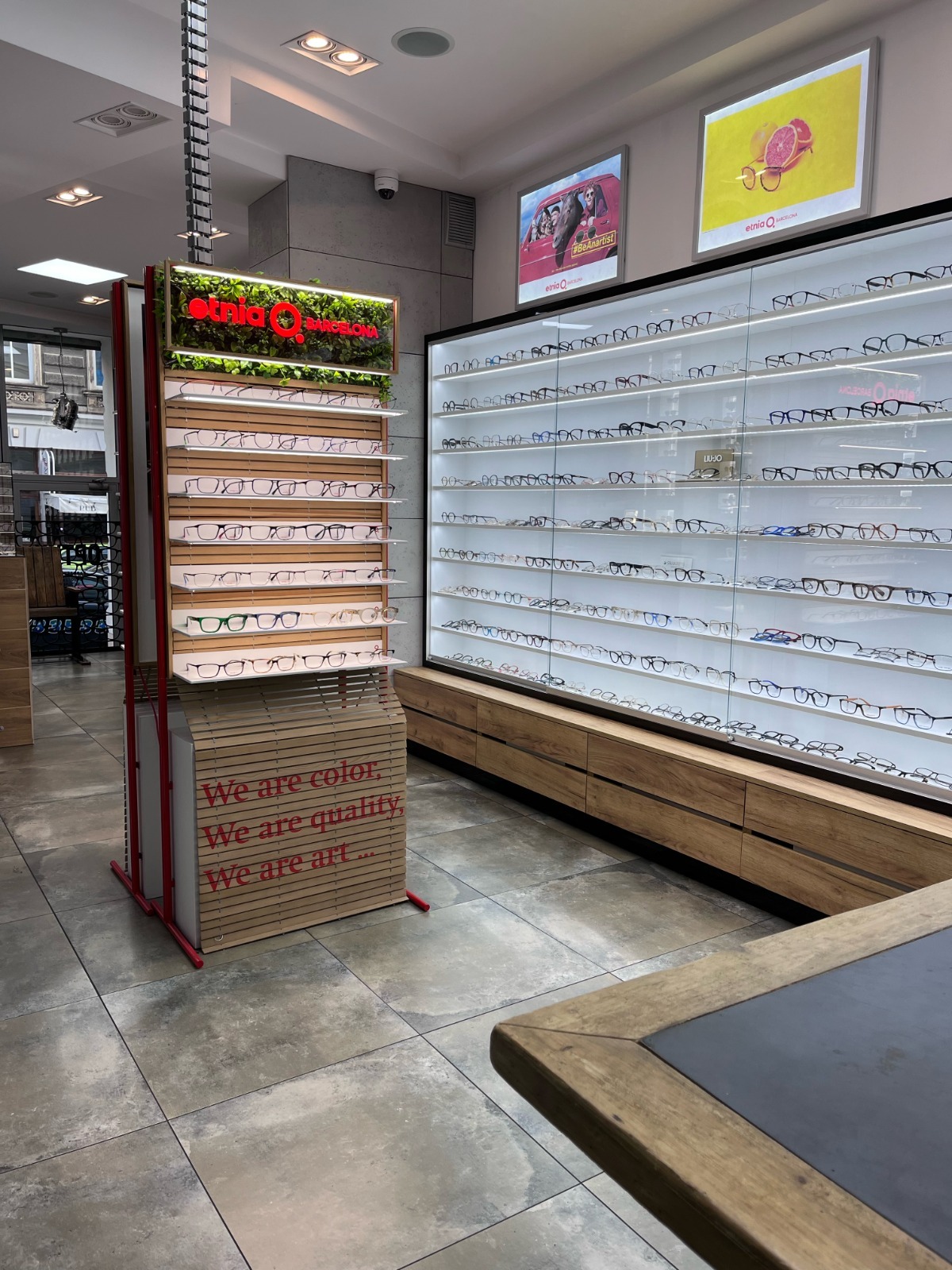 Salon optyczny Etnia Barcelona: ekspozycja okularów na regale z zielenią i podświetlanej ścianie. Napis: 'We are color, we are quality, we are art.'