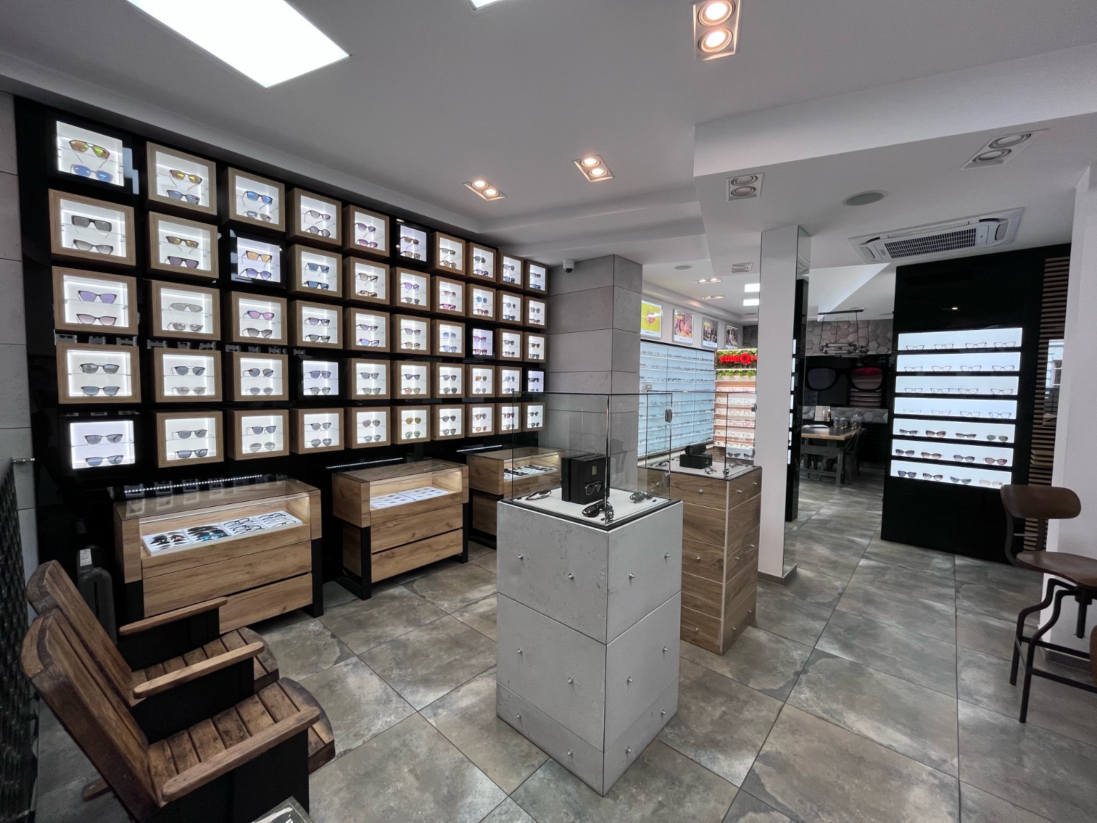 Wnętrze salonu optycznego z ekspozycją okularów w podświetlanych gablotach. Drewniane meble i szare płytki podłogowe. Nowoczesny design.
