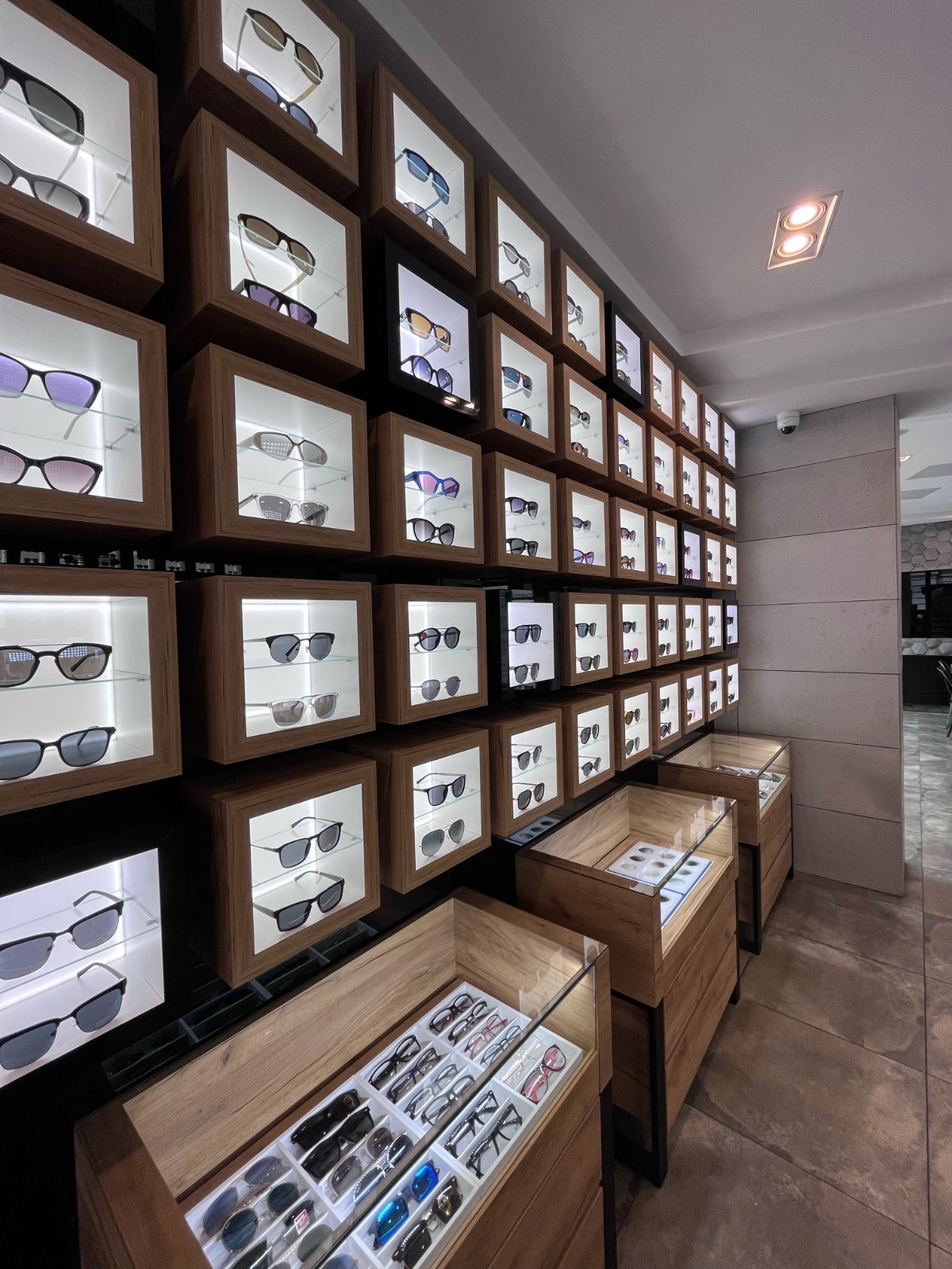 Salon optyczny: ściana z podświetlanymi gablotami okularów i ekspozytory pod szkłem. Szeroki wybór oprawek w nowoczesnym wnętrzu.