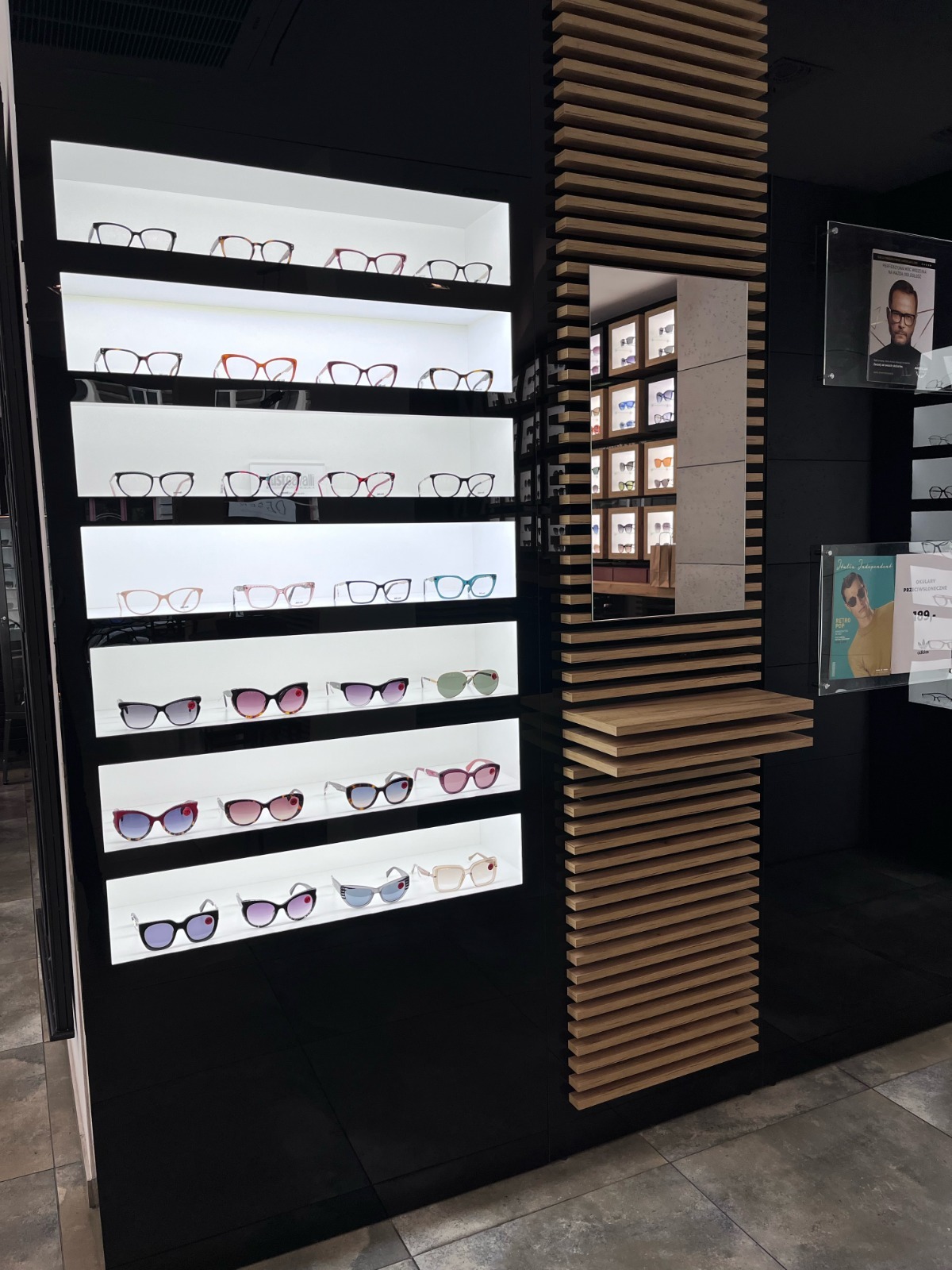 Wystawa okularów korekcyjnych i przeciwsłonecznych w salonie optycznym. Okulary ułożone na podświetlanych półkach, obok dekoracyjny element z drewnianych listewek.