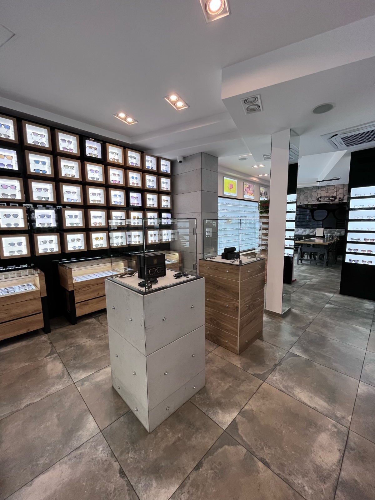 Wnętrze salonu optycznego: ekspozycja okularów w podświetlanych gablotach na ścianie i w szklanych witrynach na postumentach. Nowoczesny design, jasne oświetlenie.