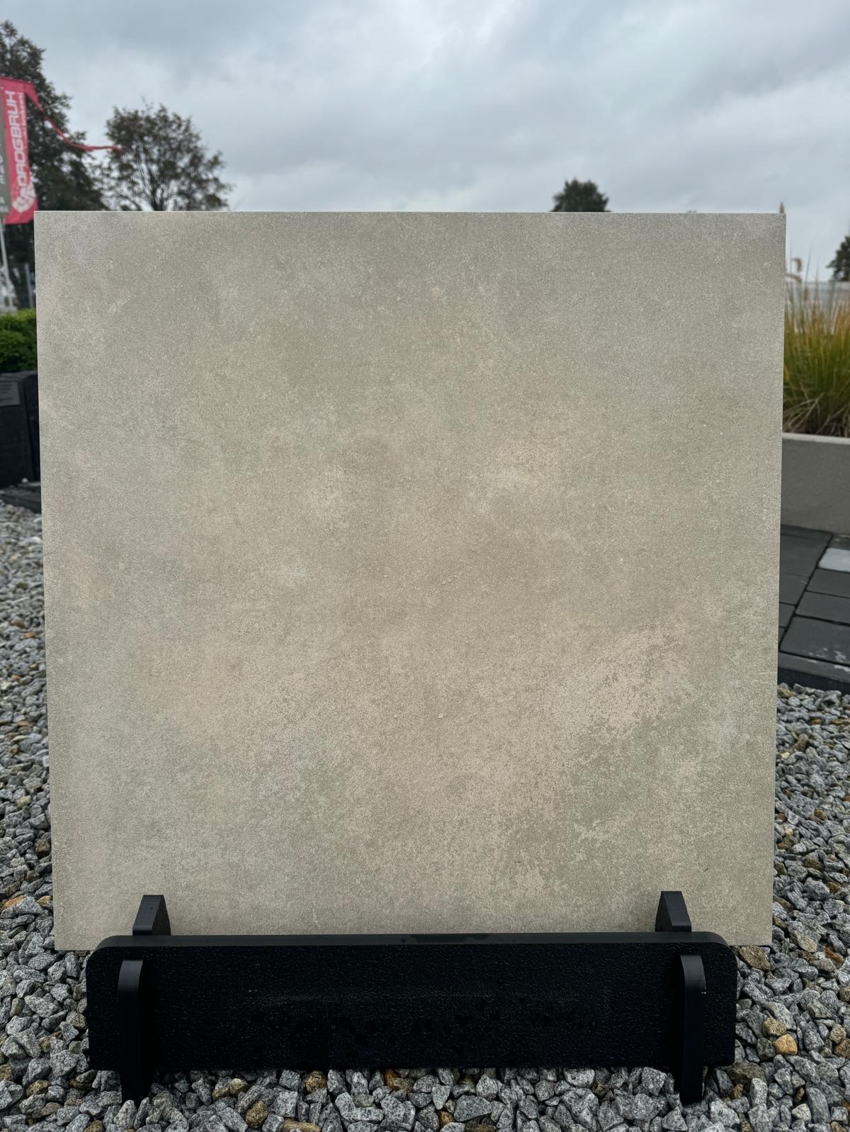 Prezentacja płyty betonowej o fakturze kamienia w odcieniach szarości, umieszczonej na czarnym stojaku, na tle żwirowego podłoża i zieleni.
