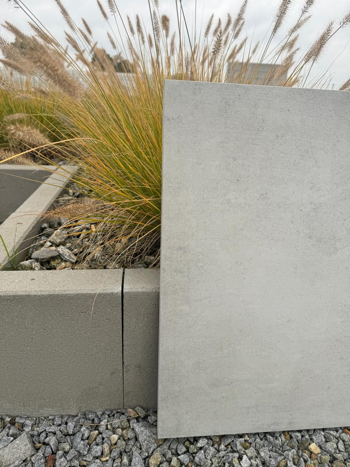 Szara płyta betonowa, prawdopodobnie element ogrodzenia, oparta o krawężnik z roślinami ozdobnymi w tle. Prezentacja materiału z bliska.
