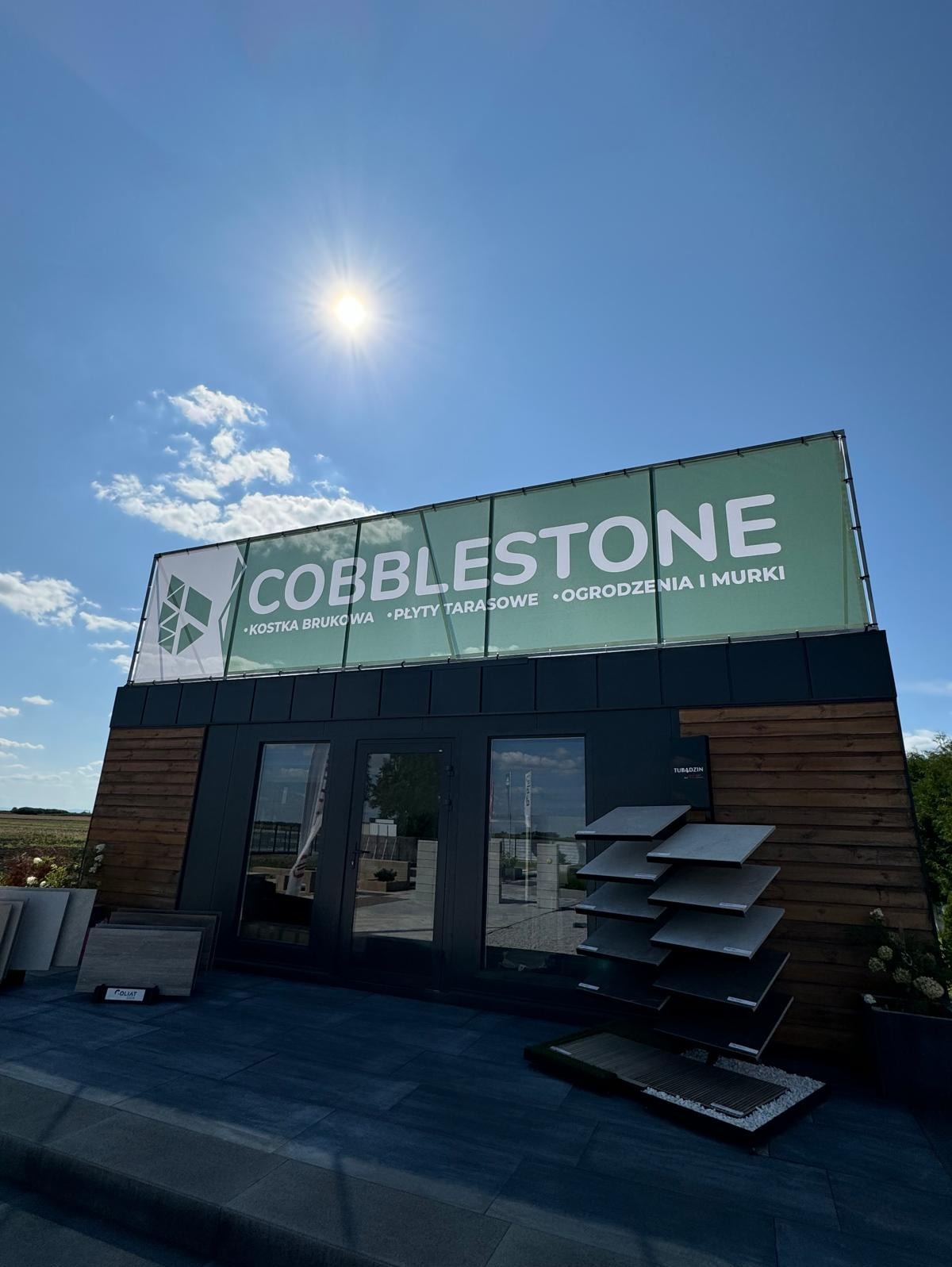 Ekspozycja kostki brukowej i płyt tarasowych marki Cobblestone. Widoczny budynek z drewnianą elewacją, baner reklamowy i próbki materiałów na zewnątrz.