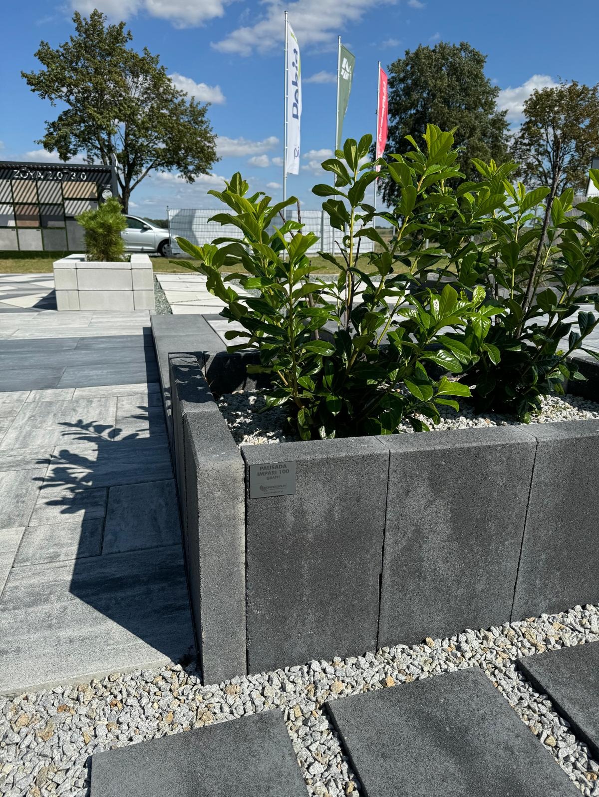 Betonowa palisada IMPARI 100 w kolorze grafitowym tworzy rabatę z zielenią na tle kostki brukowej i ekspozycji materiałów budowlanych w słoneczny dzień.