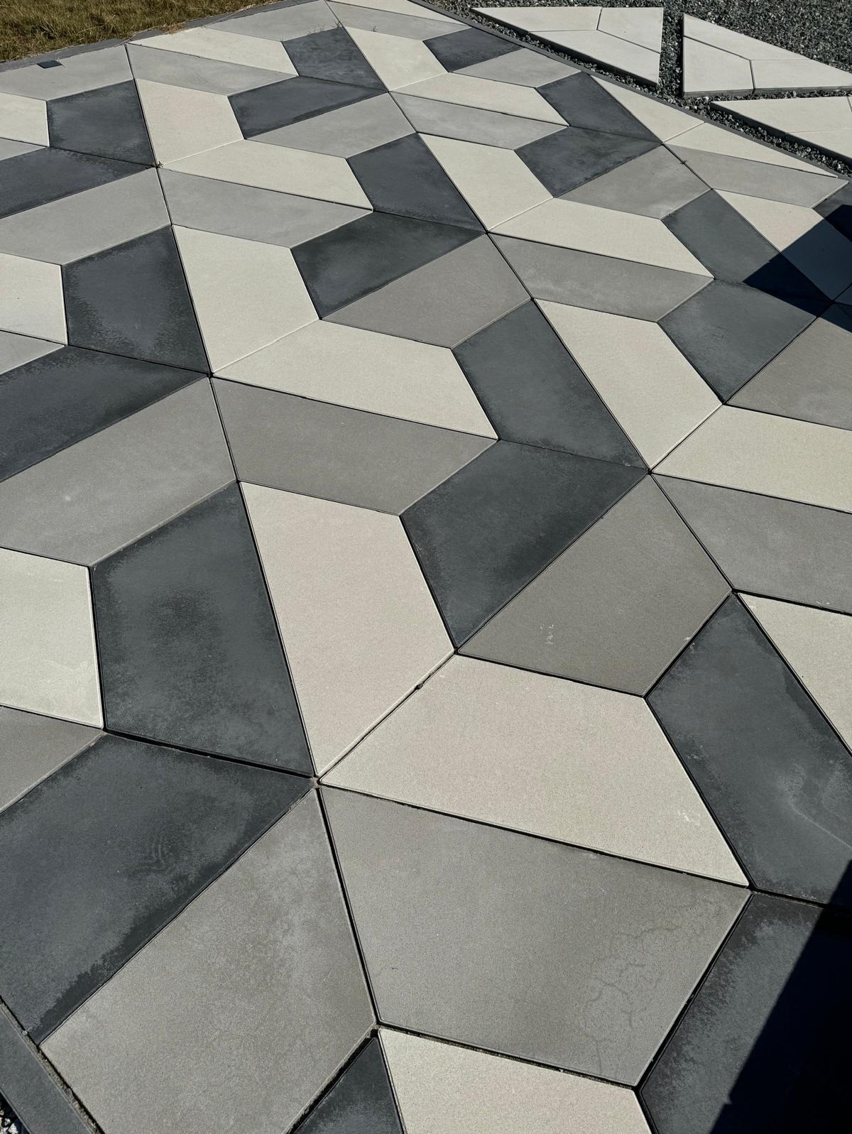 Ułożona nawierzchnia z geometrycznych płyt betonowych w odcieniach szarości, tworzących iluzję 3D. Widoczne nieułożone płyty i żwir.
