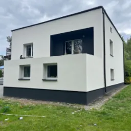 Dwukondygnacyjny dom z białą elewacją i ciemnymi akcentami, widoczny z perspektywy trawnika. Na balkonie ułożone płyty. Minimalistyczny design budynku.