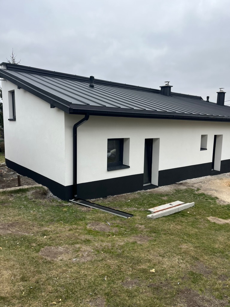 Nowoczesny, parterowy dom z białą elewacją i czarnym dachem z blachy na rąbek stojący. Widoczne rynny i fragment terenu wokół budynku.