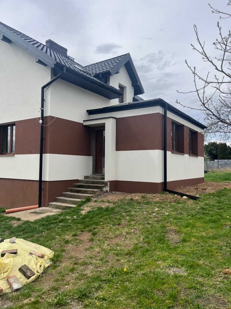 Dwukondygnacyjny dom z elewacją w tonacji bieli i brązu, widok z poziomu gruntu na front budynku z betonowymi schodami wejściowymi i elementami orynnowania.