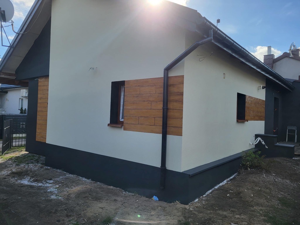 Elewacja domu jednorodzinnego po remoncie: kremowe ściany, ciemnoszary cokół i obramowania okien, panele imitujące drewno. Słoneczny dzień.