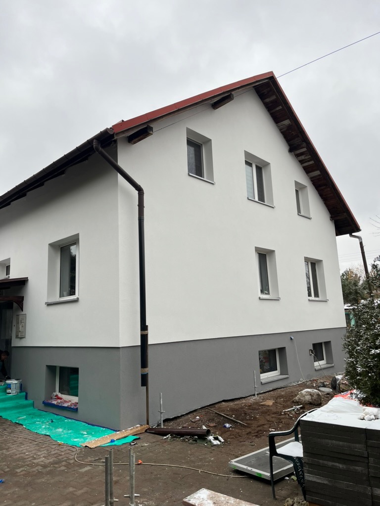 Dwukondygnacyjny dom z białą elewacją i szarym cokołem, po renowacji. Widoczne okna z białymi ramami i brązowy dach. Na ziemi resztki materiałów budowlanych.