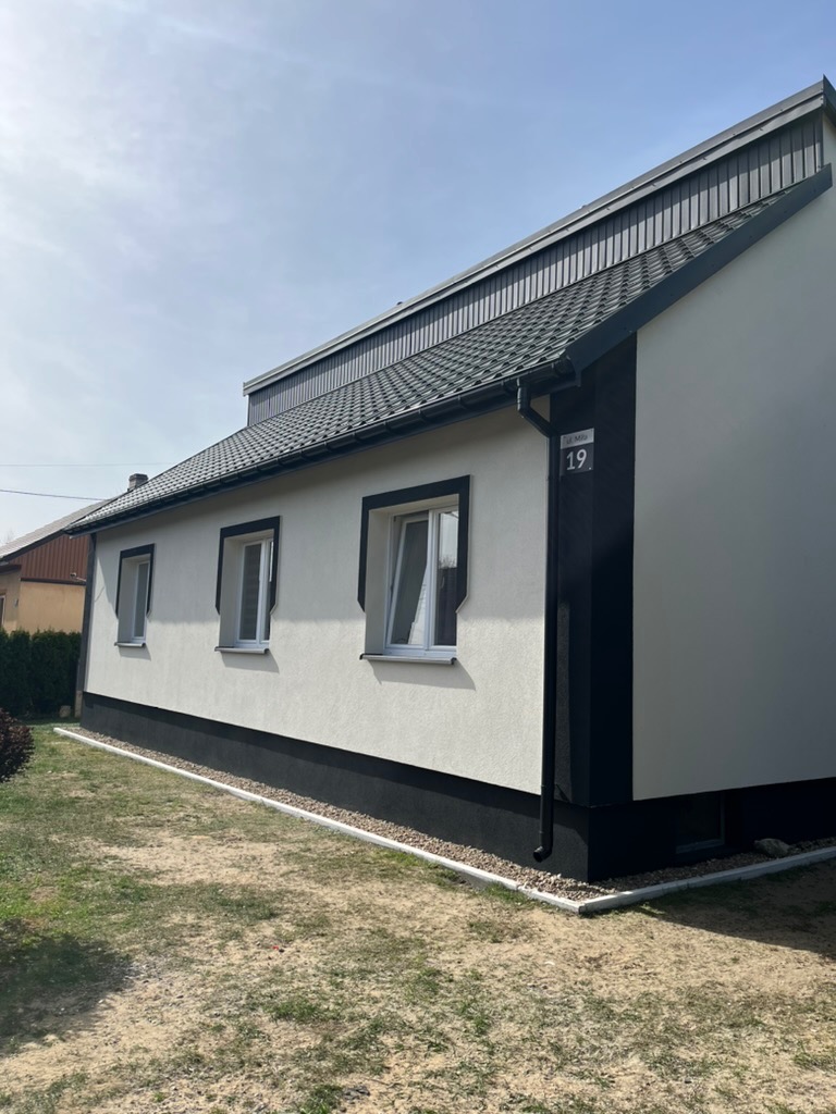Wykończony dom jednorodzinny z białą elewacją i czarnymi detalami: okna, cokół, rynna. Widoczny numer domu '19'. Ujęcie z boku, w słoneczny dzień.
