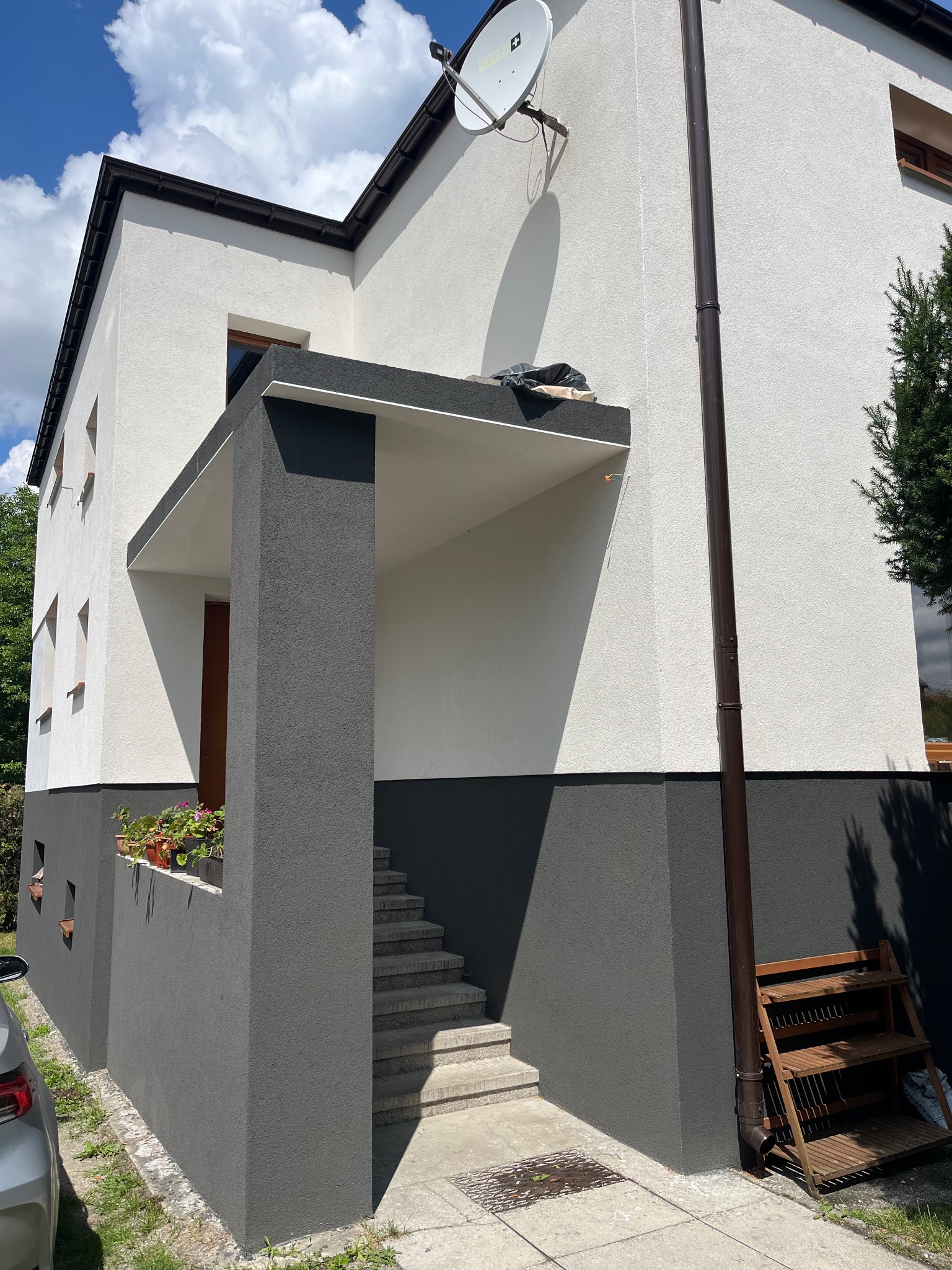 Dwukolorowa elewacja domu z szarą podstawą i białym wykończeniem, widok z boku, z anteną satelitarną i schodami wejściowymi. Słoneczny dzień.