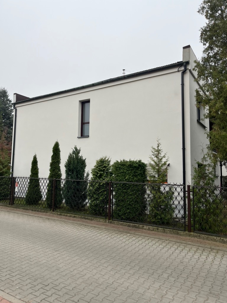 Biała, dwukondygnacyjna elewacja budynku z czarnymi akcentami, widoczna z poziomu ulicy. W tle pochmurne niebo, a przed budynkiem ozdobne krzewy i metalowe ogrodzenie.