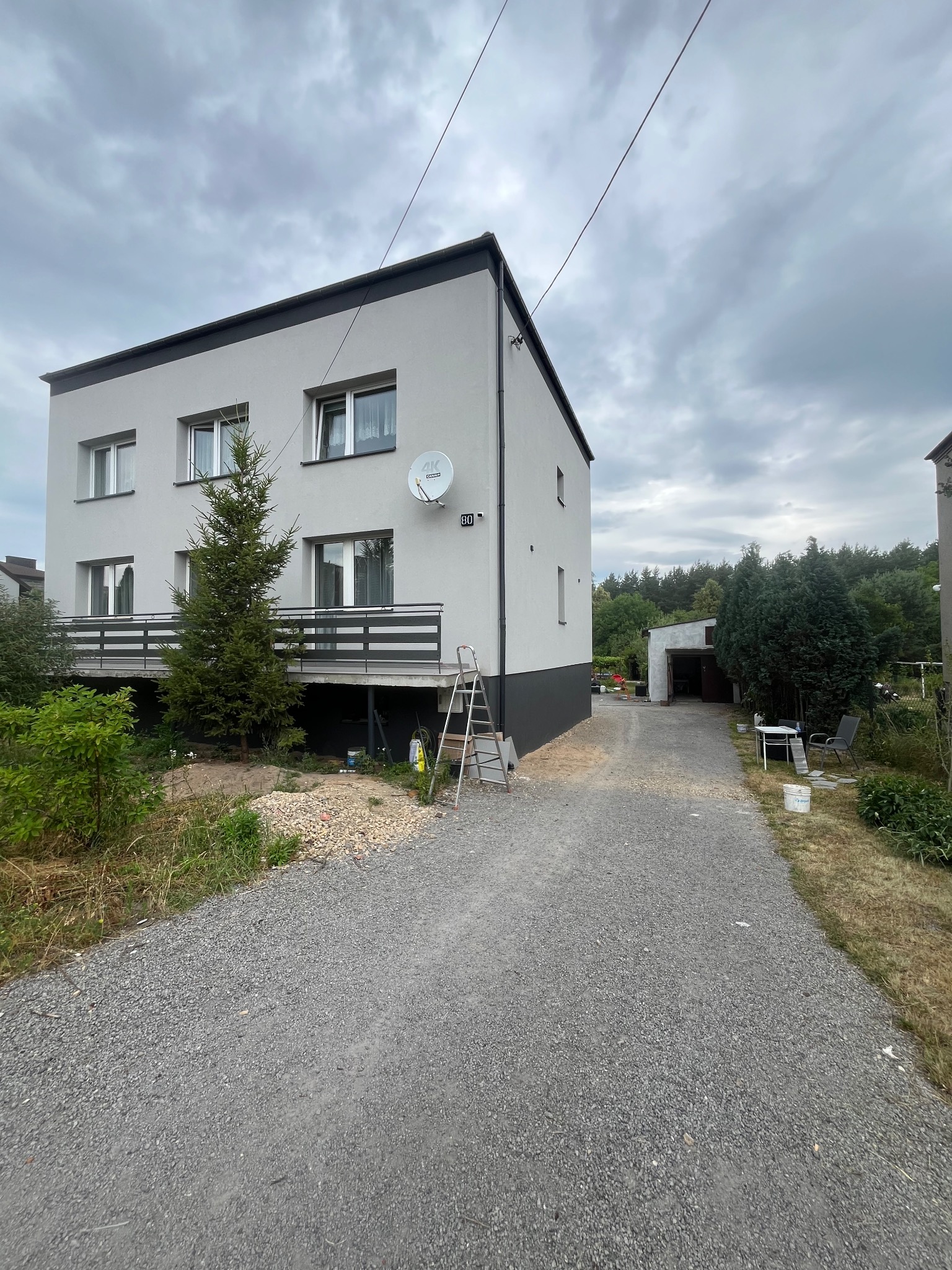 Dwukondygnacyjny dom z szarą elewacją i ciemnoszarym cokołem. Widoczna drabina oparta o ścianę, sugerująca prace elewacyjne w toku. Na balkonie rośnie choinka.