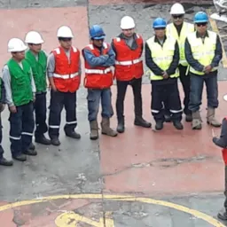 Szkolenie BHP na budowie: grupa pracowników w kaskach i kamizelkach odblaskowych słucha instruktora. Widok z góry na plac z żółtym symbolem na czerwonym tle.