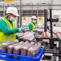 JOBHORA SPÓŁKA Z OGRANICZONĄ ODPOWIEDZIALNOŚCIĄ - Pracownicy magazynu w kaskach i maskach skanują zapakowane produkty. Widoczne regały, stanowiska pracy i oznaczenia numeracyjne. Hala produkcyjna.