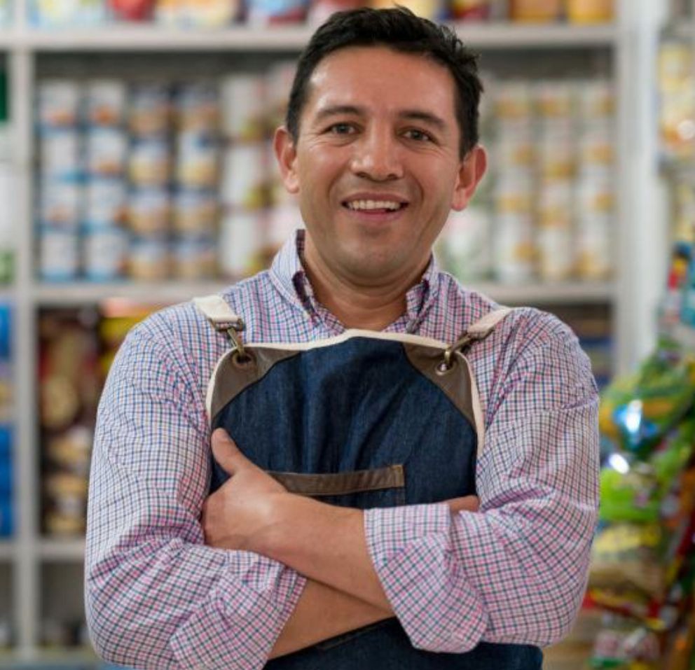 Uśmiechnięty mężczyzna w dżinsowym fartuchu, z założonymi rękami na tle regałów sklepowych. Pracownik sklepu spożywczego prezentuje się w miejscu pracy.