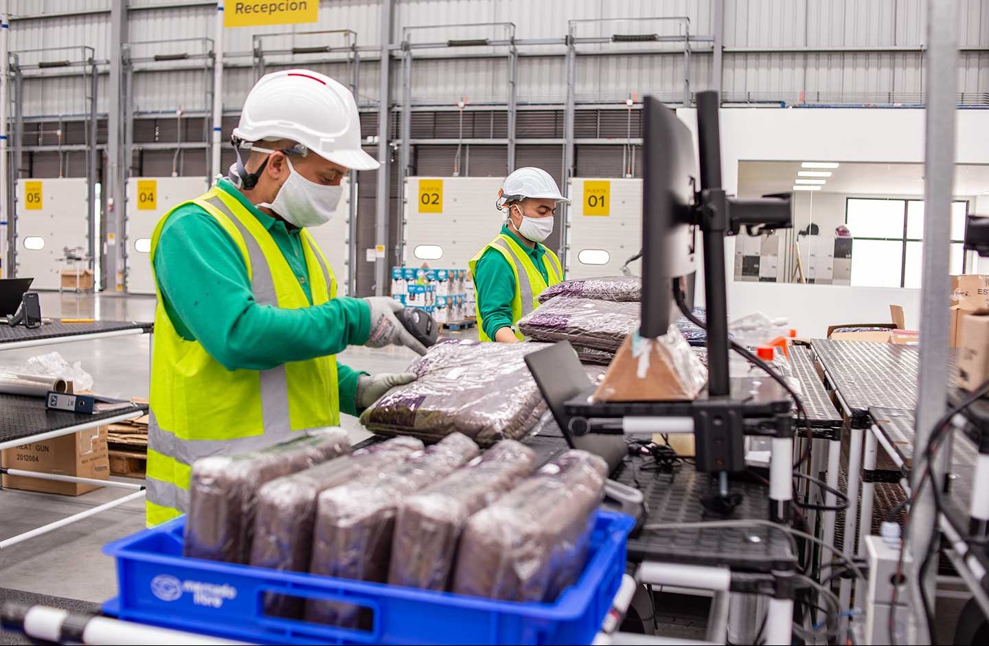 Pracownicy magazynu w kaskach i maskach skanują zapakowane produkty. Widoczne regały, stanowiska pracy i oznaczenia numeracyjne. Hala produkcyjna.