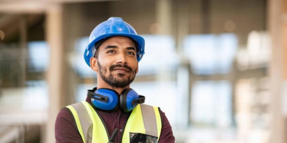 Uśmiechnięty pracownik budowlany w kasku, kamizelce odblaskowej i słuchawkach ochronnych na tle rozmytej konstrukcji budynku.