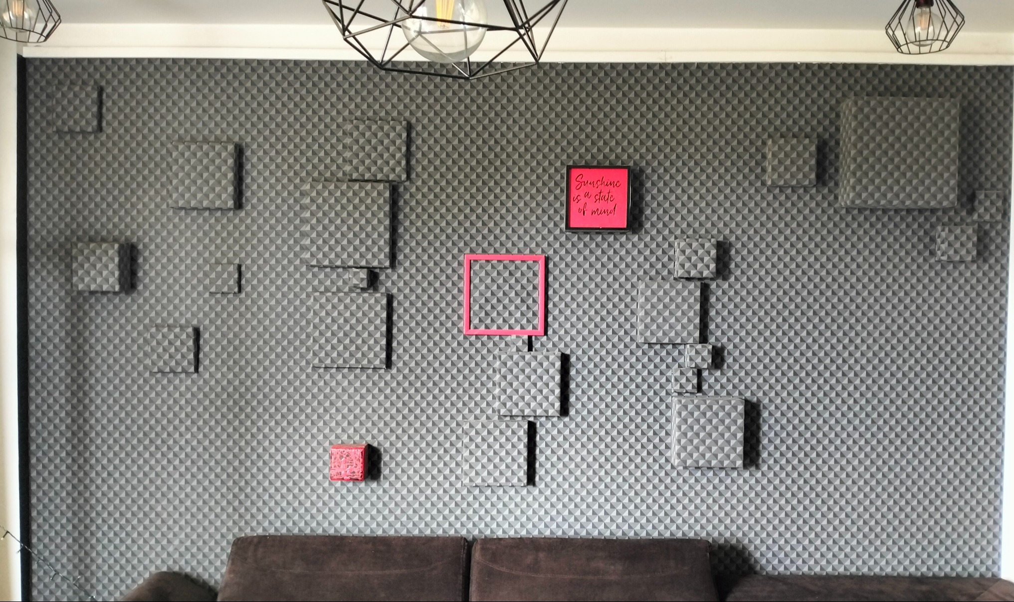Szara ściana z geometrycznym wzorem i wystającymi kwadratami, urozmaicona różowymi akcentami: ramką i małym pudełkiem. Widoczny fragment sofy i nowoczesne lampy.