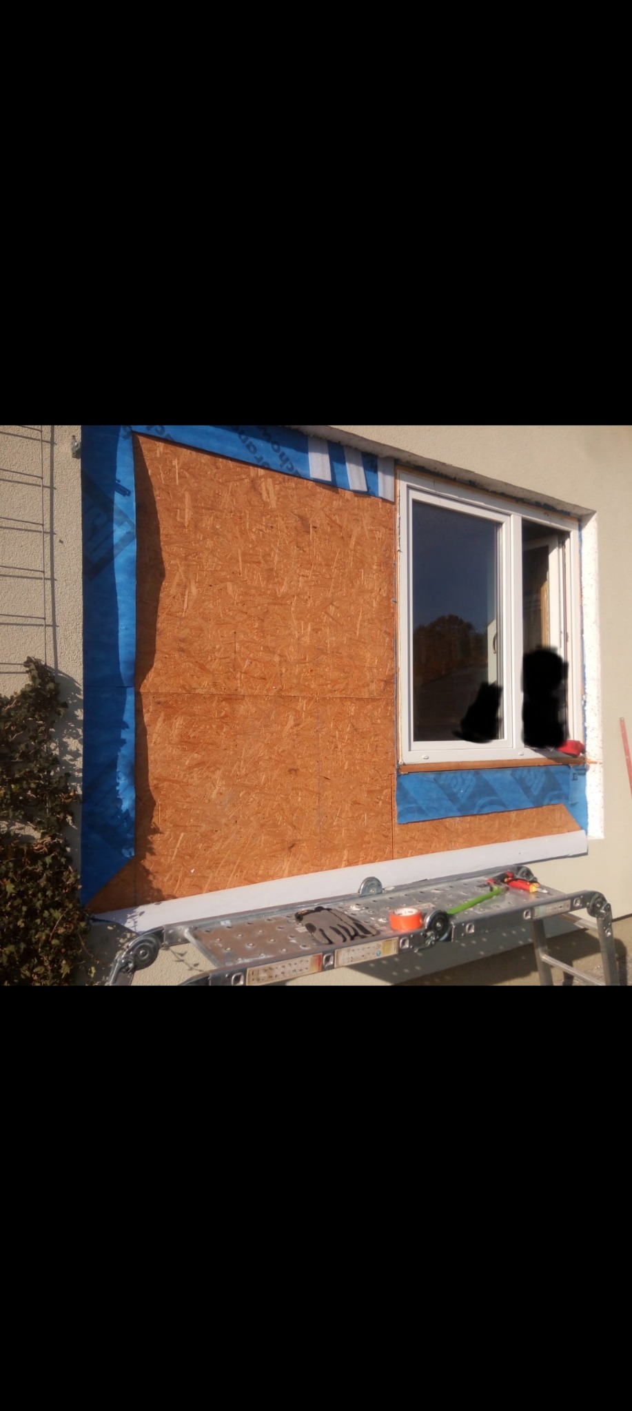 Przygotowanie do montażu okna: płyta OSB zabezpieczona folią, rusztowanie z narzędziami, widok z zewnątrz na ścianę budynku.