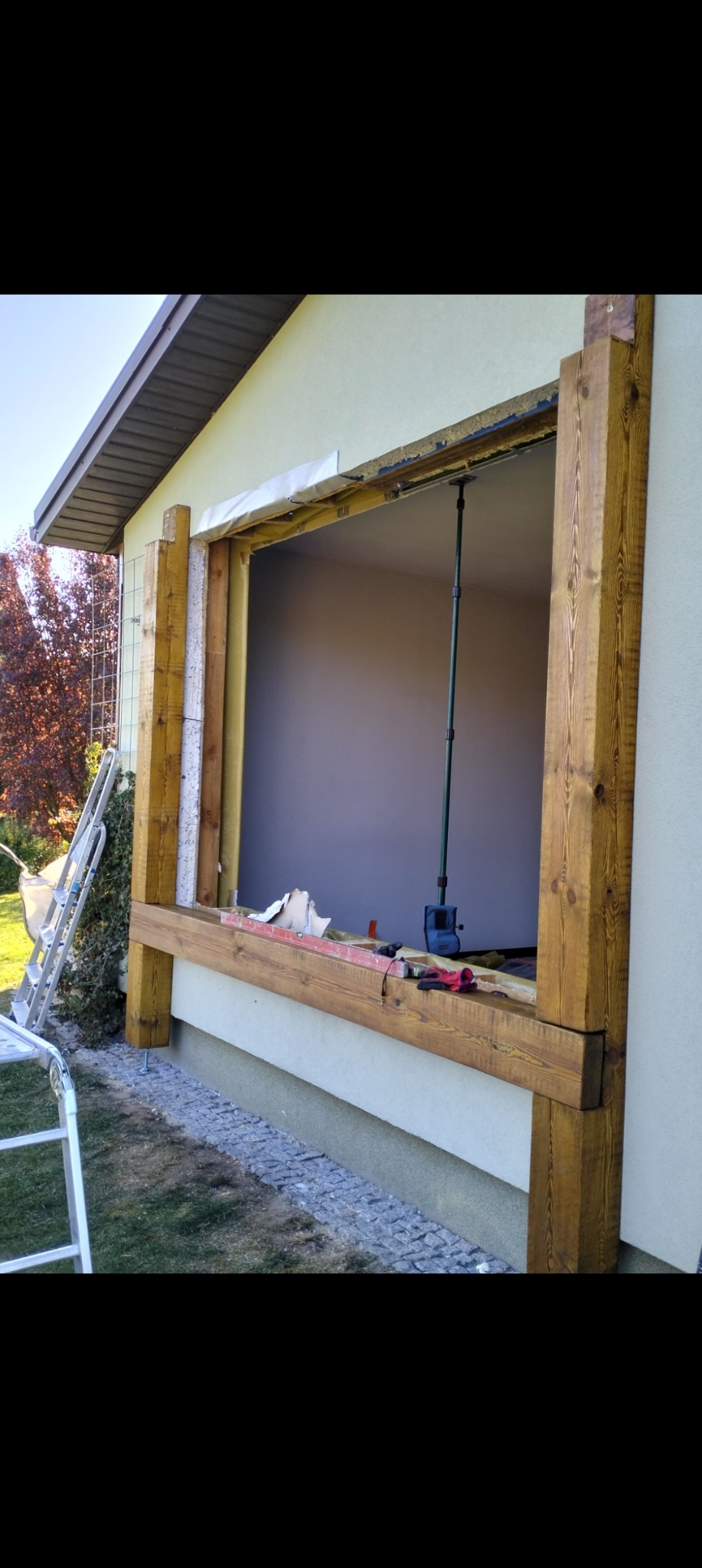 Montaż drewnianej ramy okiennej w budynku z ociepleniem, widoczne narzędzia i drabina. Prace w toku, z zewnątrz budynku.