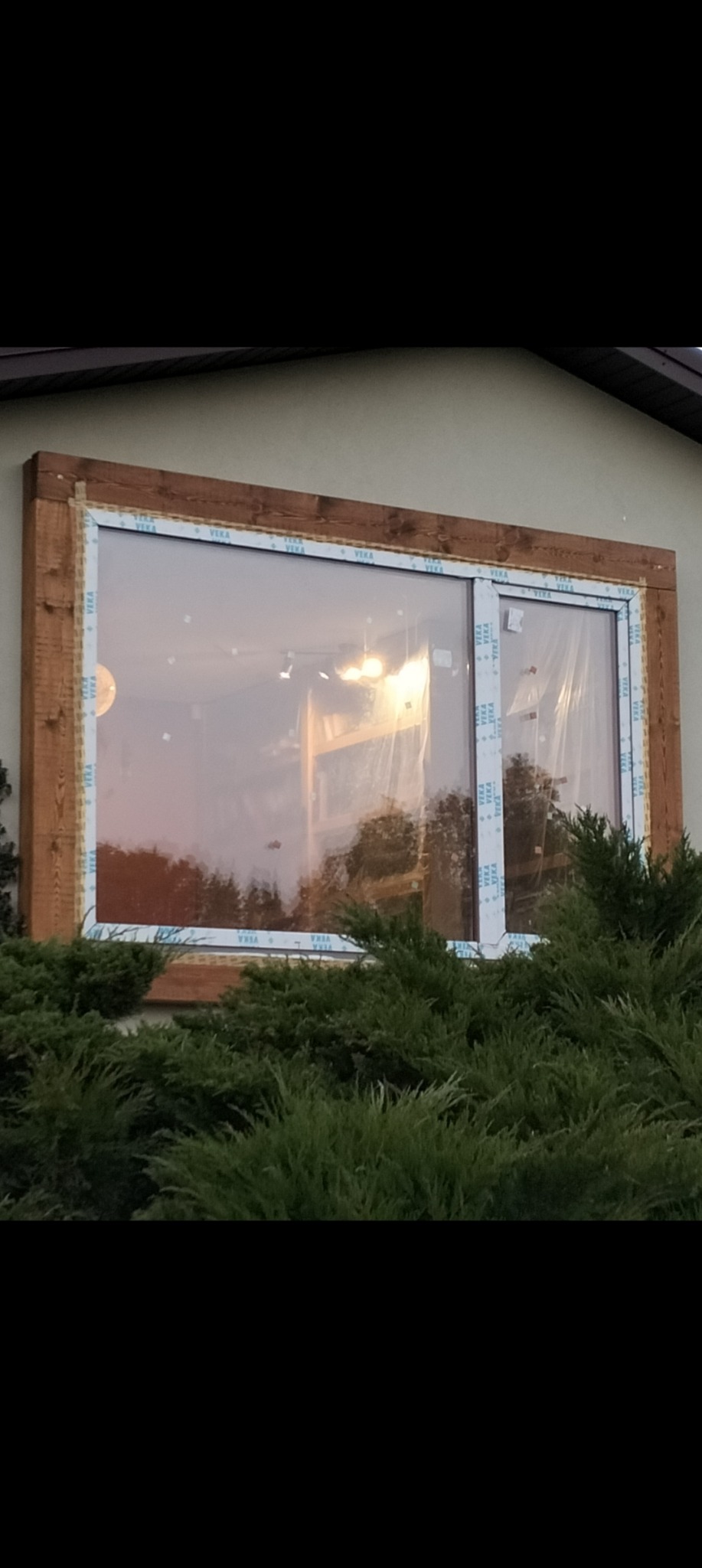 Nowe, dwuskrzydłowe okno PCV z drewnianą ramą montażową, widoczne z zewnątrz budynku, częściowo zasłonięte krzewami, w trakcie montażu.