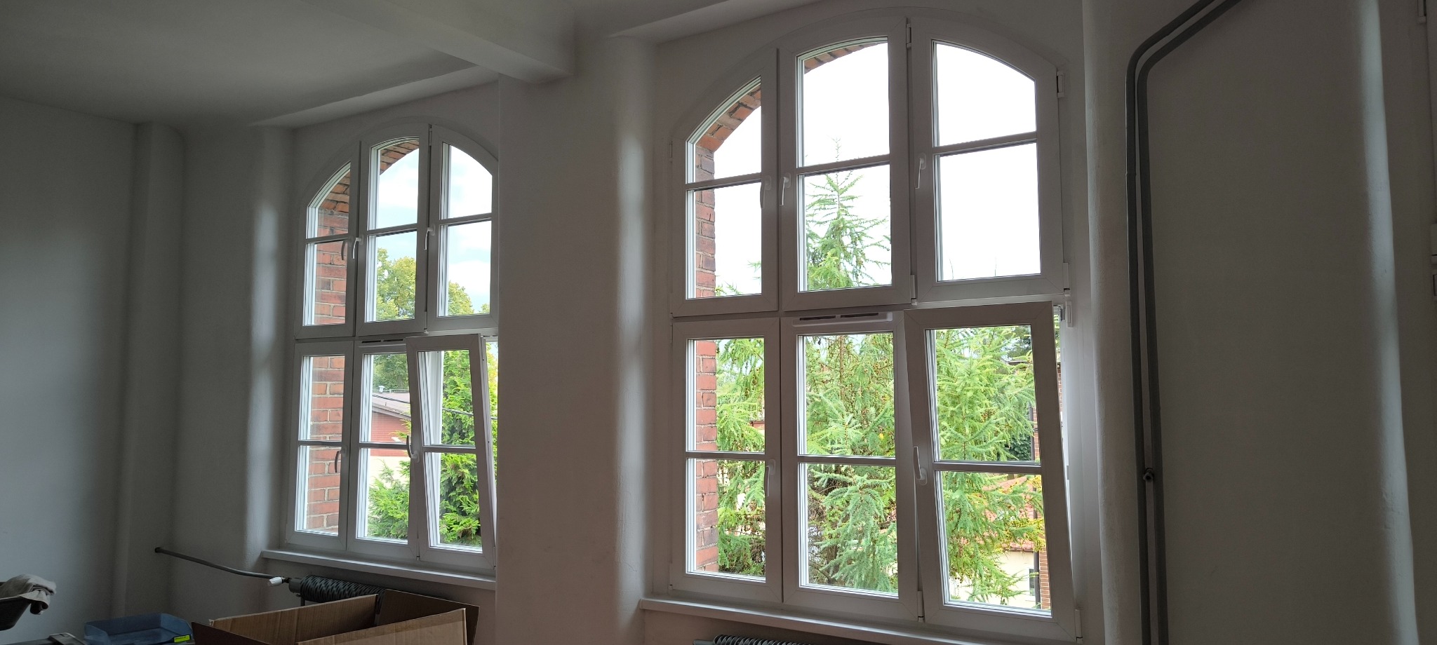 Dwa okna z białymi ramami, jedno otwarte, z widokiem na zieleń. Widoczne grzejniki i kartonowe pudła na podłodze, co sugeruje przeprowadzkę lub remont.
