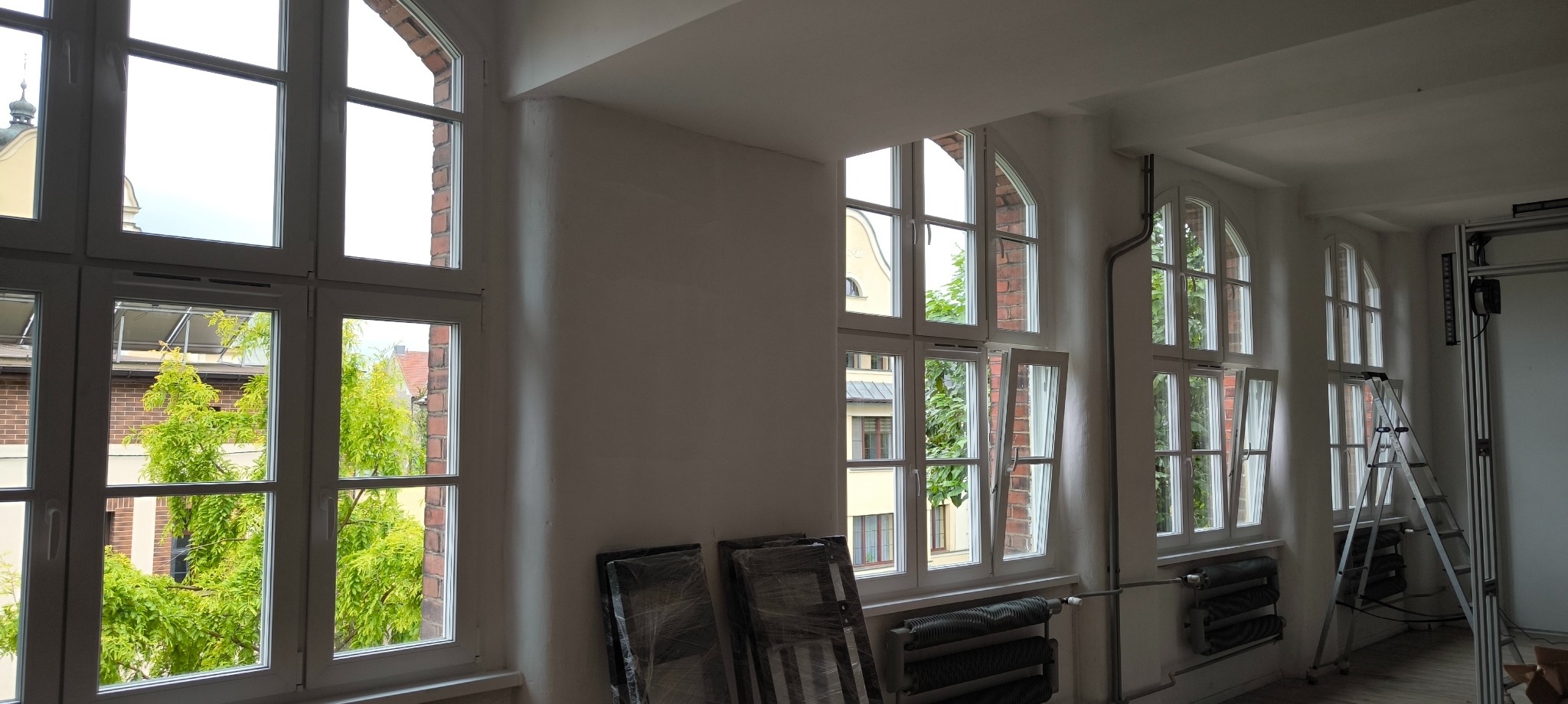 Wnętrze z rzędem białych, łukowych okien. Część okien otwarta, widok na zieleń i budynki. Aluminiowa drabina i grzejniki pod oknami. Folią zabezpieczone elementy wyposażenia.