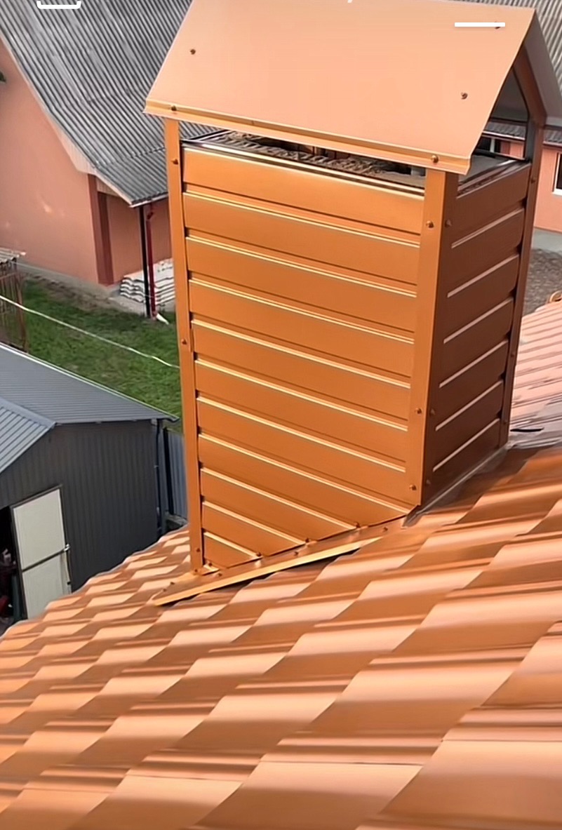 Nowy, miedziany komin wentylacyjny z blachy na dachu z blachodachówki w kolorze miedzi, widok z góry. W tle budynki mieszkalne i zieleń.