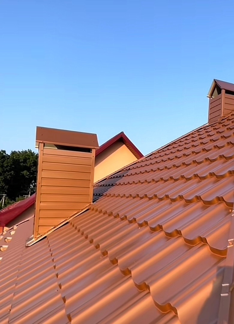 Brązowy dach z blachy trapezowej i kominy wentylacyjne na tle błękitnego nieba w słoneczny dzień. Widok z dołu.