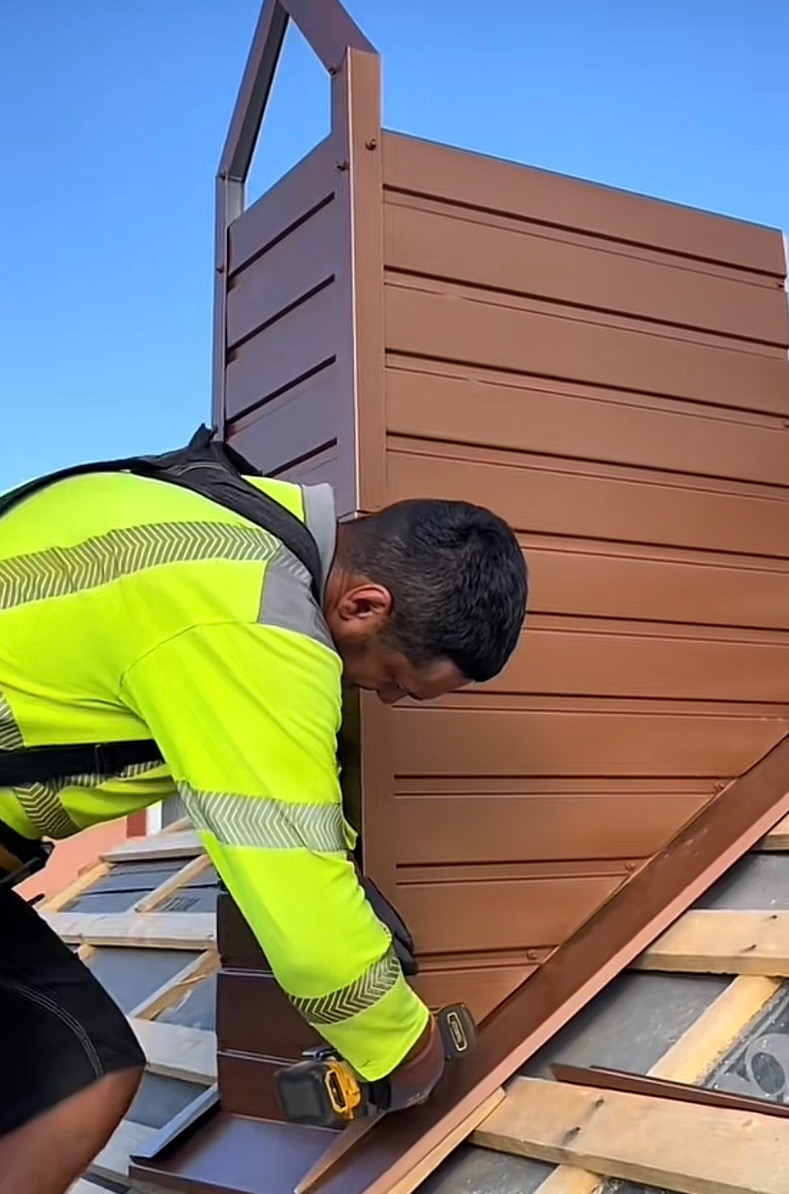 Dekarz w żółtej kamizelce montuje brązowe obróbki blacharskie na dachu, używając wkrętarki. Widoczne dachówki i fragment komina.