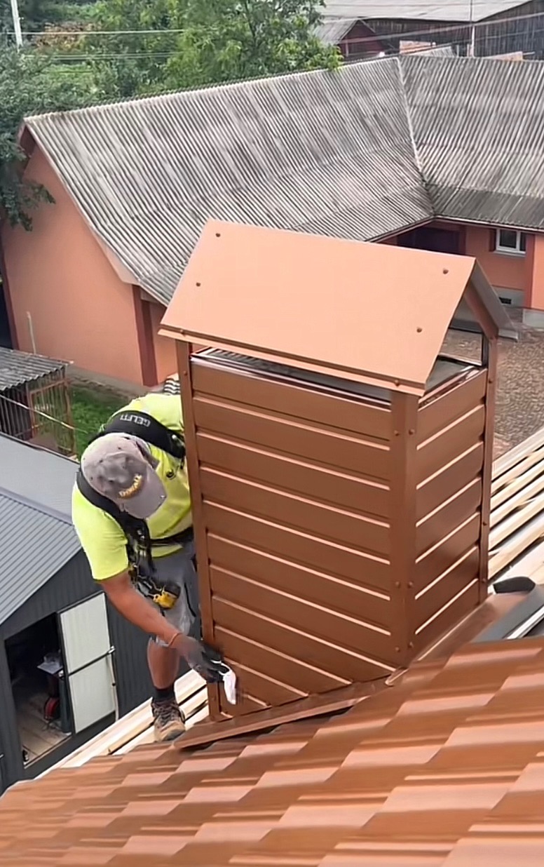 Dekarz montuje brązową obróbkę blacharską komina na dachu z blachodachówki w słoneczny dzień, widok z góry.