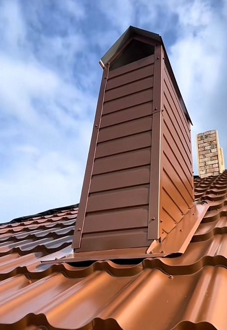 Brązowy komin z blachy na dachu z blachodachówki w kolorze miedzianym, widoczny drugi komin w tle, błękitne niebo.