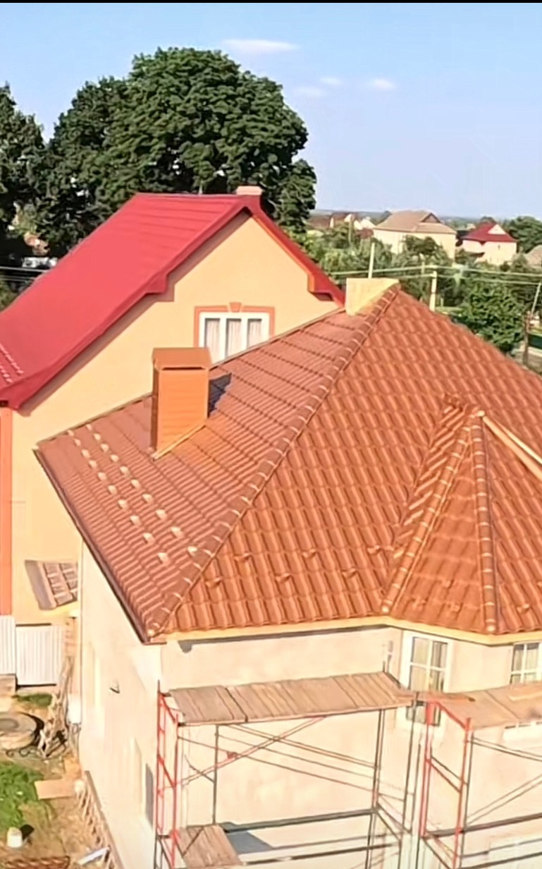 Nowy, brązowy dach z dachówki ceramicznej na jasnym domu, widoczny z góry, z rusztowaniem w tle. Drugi dom z czerwonym dachem w oddali.
